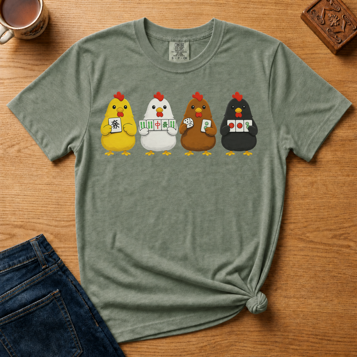 Mahjong Chickens Shirt in Moss - four cartoon hens holding mahjong tiles on a green Comfort Colors 1717 tee, flat lay