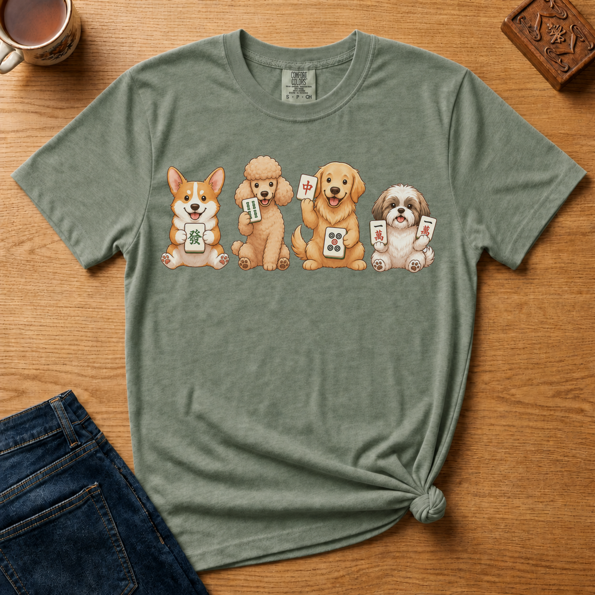 Mahjong Dogs Shirt in Moss - four cartoon dogs holding mahjong tiles on a green Comfort Colors 1717 tee, flat lay