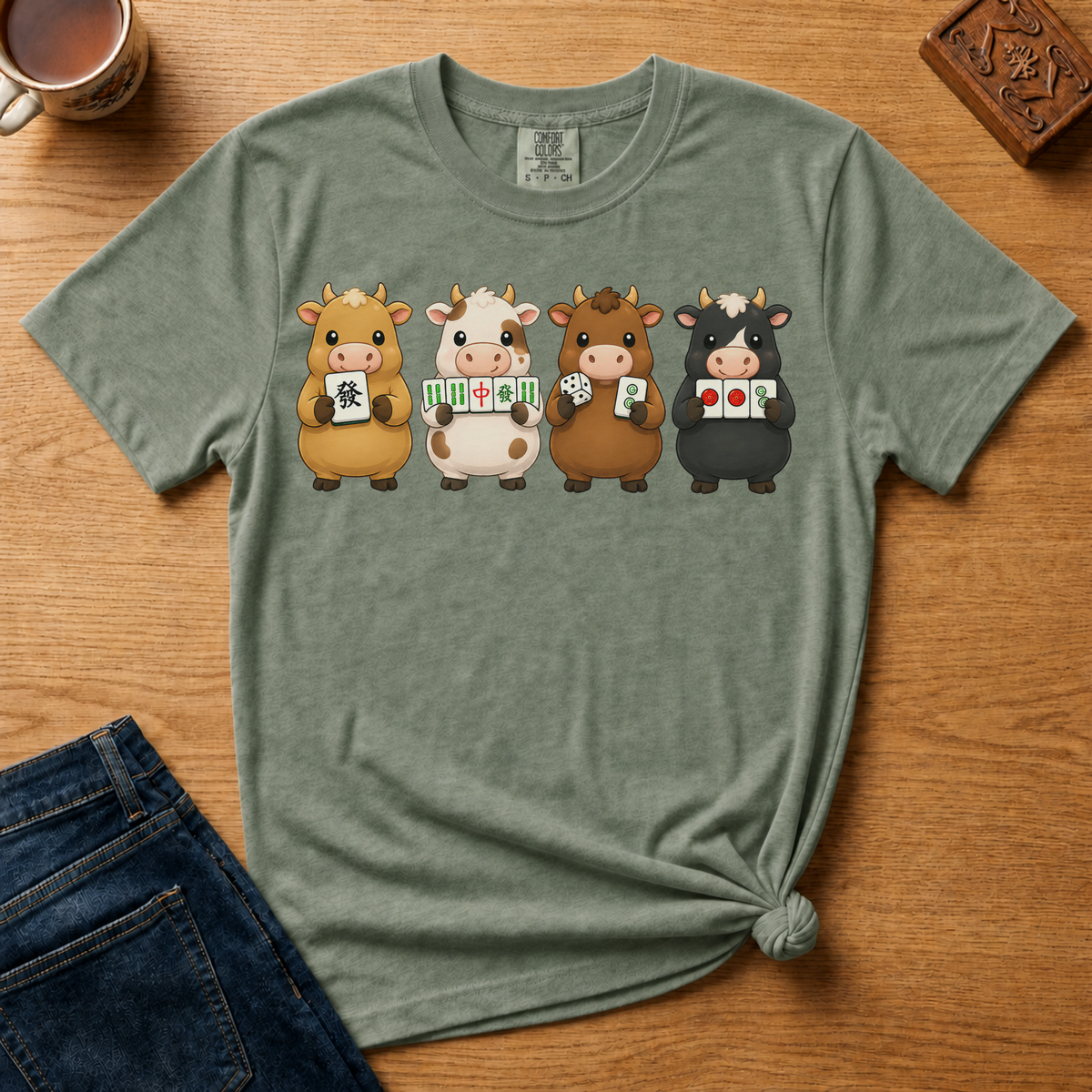 Mahjong Cows Shirt in Moss - four cartoon cows holding mahjong tiles on a green Comfort Colors 1717 tee, flat lay