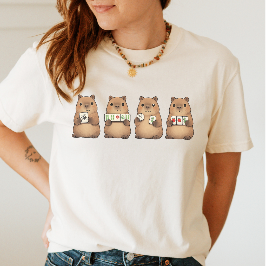 Mahjong Capybara Shirt in Ivory - four cartoon capybaras holding mahjong tiles on an ivory Comfort Colors 1717 tee, flat lay