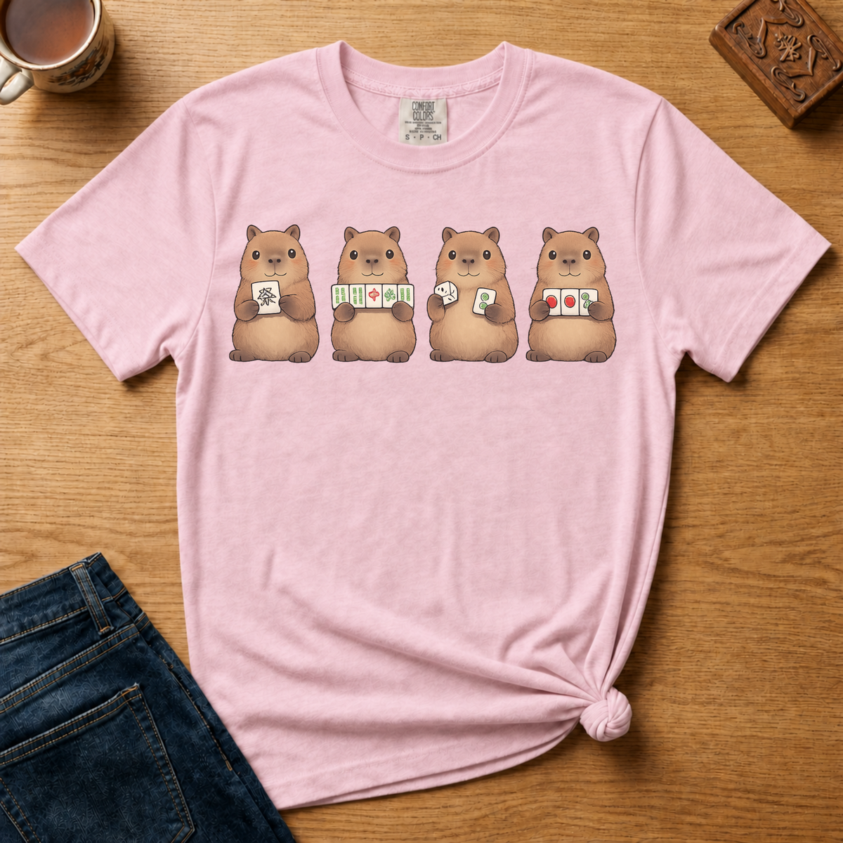 Mahjong Capybara Shirt in Black - four cartoon capybaras holding mahjong tiles on a black Comfort Colors 1717 tee, flat lay
