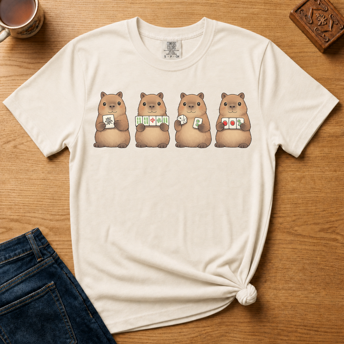 Mahjong Capybara Shirt in White - four cartoon capybaras holding mahjong tiles on a white Comfort Colors 1717 tee, flat lay
