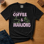She Runs On Coffee And Mahjong Shirt in Black - pink bubble lettering with coffee cup and tile doodles on a black Comfort Colors 1717 tee, flat lay