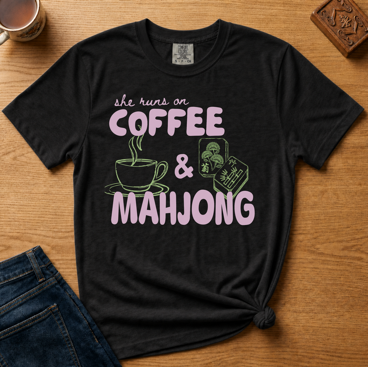She Runs On Coffee And Mahjong Shirt in Black - pink bubble lettering with coffee cup and tile doodles on a black Comfort Colors 1717 tee, flat lay