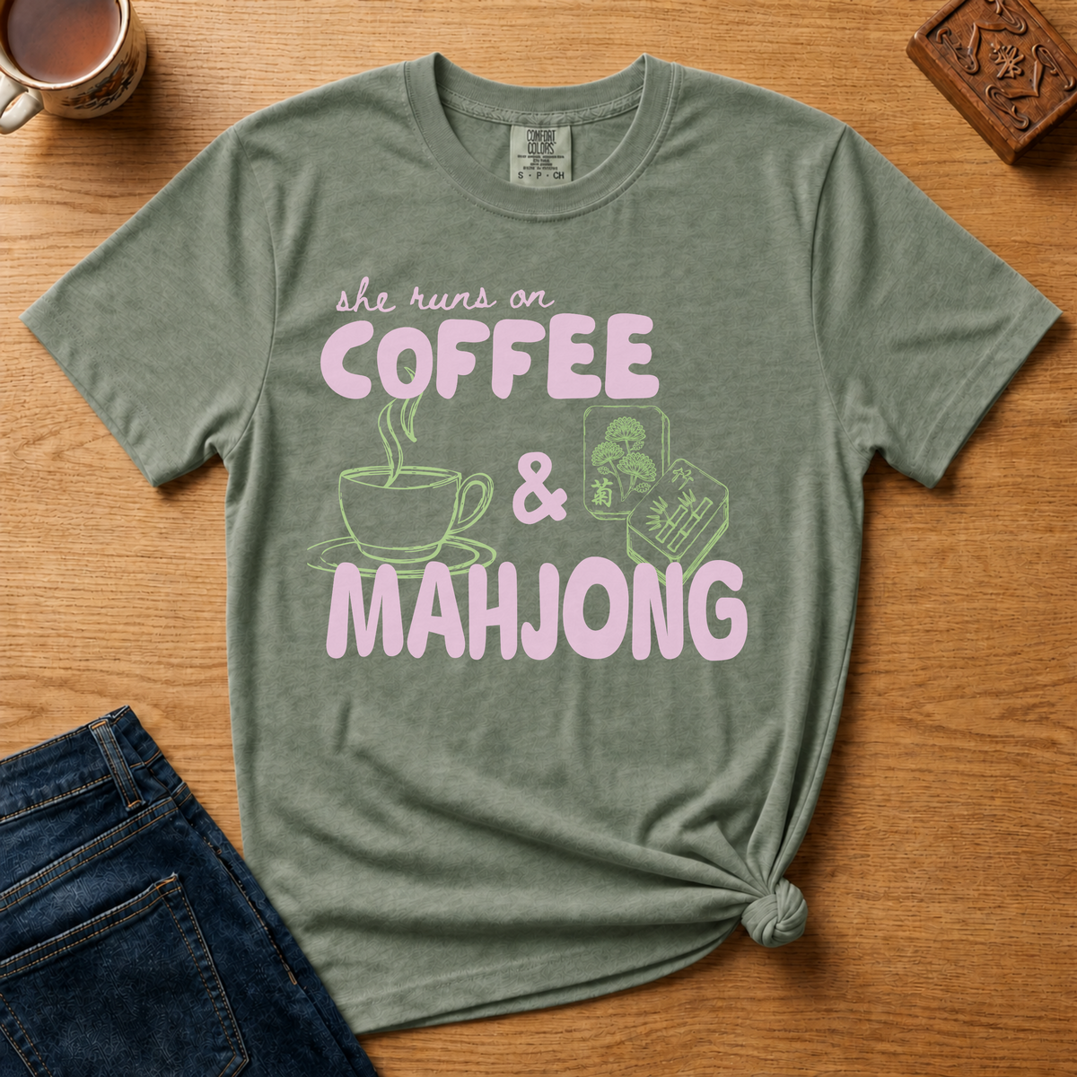 She Runs On Coffee And Mahjong Shirt in Moss - pink bubble lettering with coffee cup and tile doodles on a green Comfort Colors 1717 tee, flat lay
