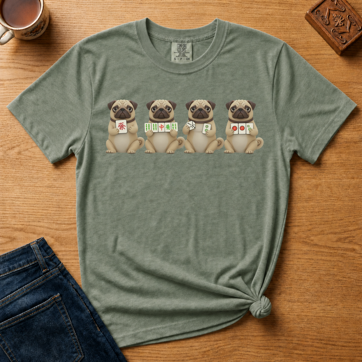 Mahjong Pugs Shirt in Moss - four cartoon pugs holding mahjong tiles on a green Comfort Colors 1717 tee, flat lay