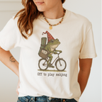Off To Play Mahjong Shirt in White - storybook frog in a polka-dot hat riding a bicycle with mahjong tiles on a white Comfort Colors 1717 tee, flat lay