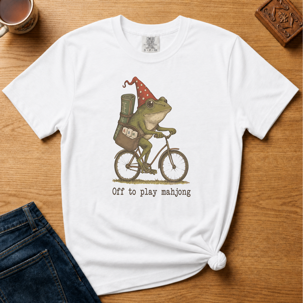 Off To Play Mahjong Shirt in Blossom - storybook frog in a polka-dot hat riding a bicycle with mahjong tiles on a pink Comfort Colors 1717 tee, flat lay
