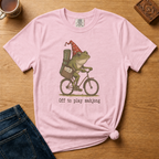 Off To Play Mahjong Shirt in Violet - storybook frog in a polka-dot hat riding a bicycle with mahjong tiles on a purple Comfort Colors 1717 tee, flat lay