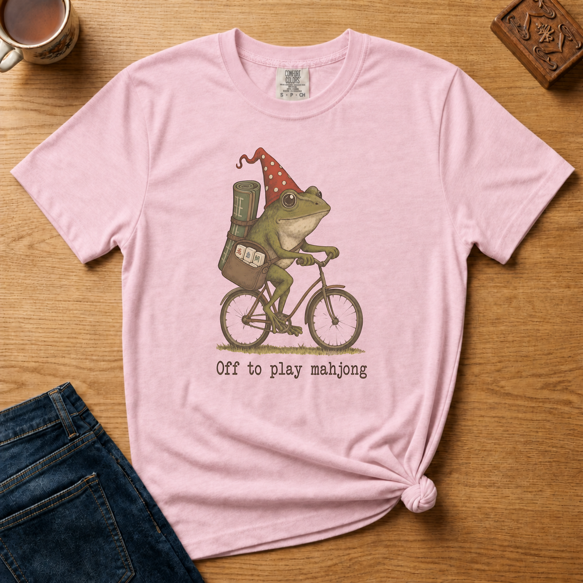 Off To Play Mahjong Shirt in Violet - storybook frog in a polka-dot hat riding a bicycle with mahjong tiles on a purple Comfort Colors 1717 tee, flat lay