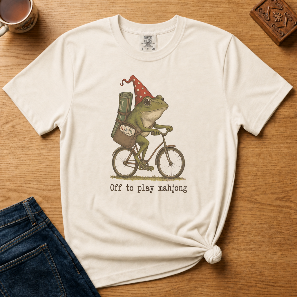 Off To Play Mahjong Shirt in Ivory - storybook frog in a polka-dot hat riding a bicycle with mahjong tiles on an ivory Comfort Colors 1717 tee, flat lay