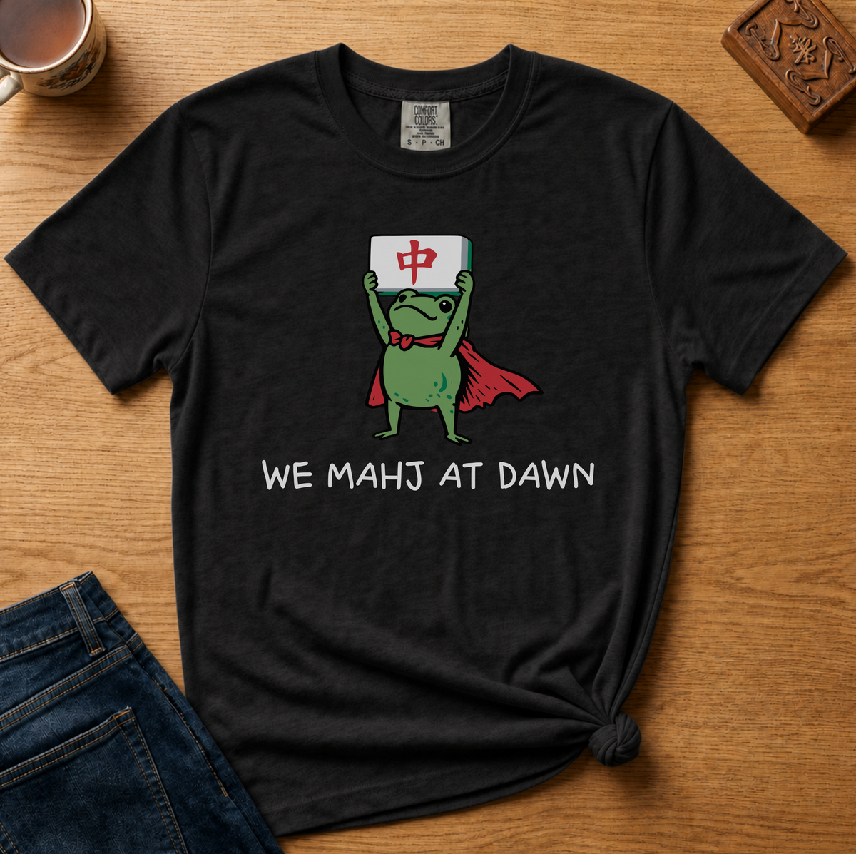 We Mahj At Dawn Shirt in Espresso - superhero frog in a red cape holding a red dragon tile overhead on a brown Comfort Colors 1717 tee, flat lay