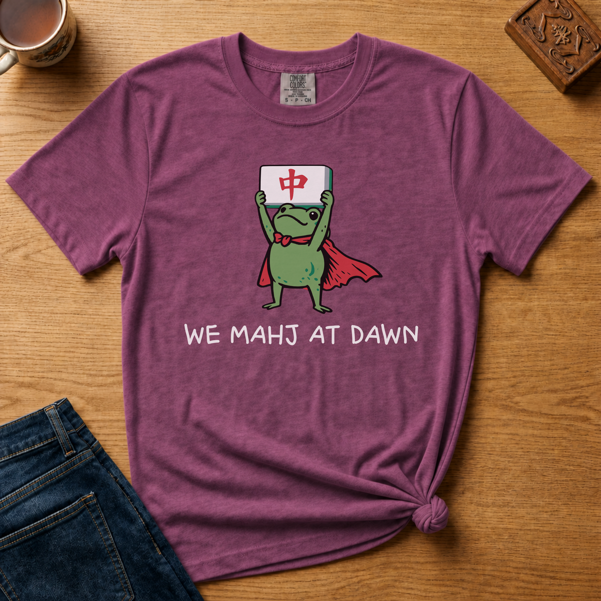 We Mahj At Dawn Shirt in Moss - superhero frog in a red cape holding a red dragon tile overhead on a green Comfort Colors 1717 tee, flat lay