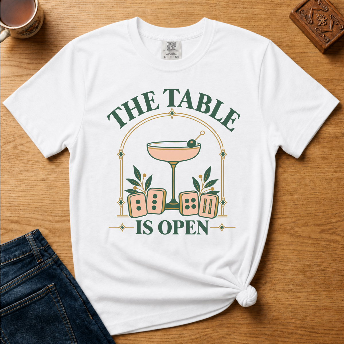 The Table Is Open Shirt in White - green serif lettering, gold deco arch, cocktail coupe and peach mahjong tiles