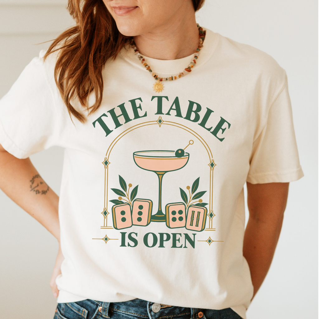 The Table Is Open Shirt in Blossom - green serif lettering, gold deco arch, cocktail coupe and peach mahjong tiles