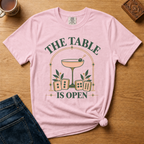 The Table Is Open Shirt in Violet - green serif lettering, gold deco arch, cocktail coupe and peach mahjong tiles