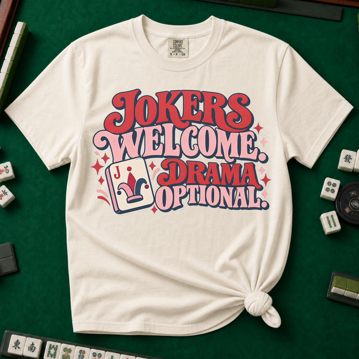 Jokers Welcome Drama Optional T-Shirt in Ivory - tee laid flat on a green mahjong mat with scattered tiles, printed with the words Jokers Welcome Drama Optional in retro bubble lettering with a joker tile