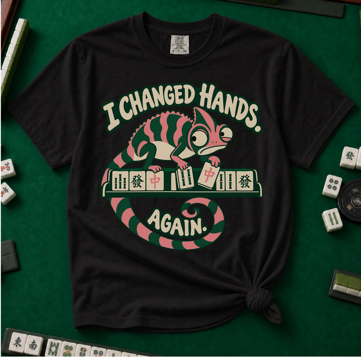 I Changed Hands Again T-Shirt in Ivory - knotted tee on a green mahjong mat with scattered tiles, printed with a chameleon standing on a rack of mahjong tiles holding a red dragon tile, and the words I Changed Hands Again