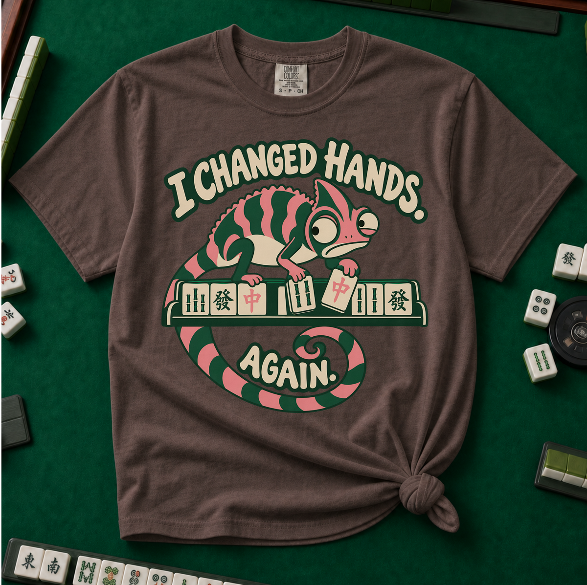 I Changed Hands Again T-Shirt in Espresso - knotted tee on a green mahjong mat with scattered tiles, printed with a chameleon standing on a rack of mahjong tiles holding a red dragon tile, and the words I Changed Hands Again