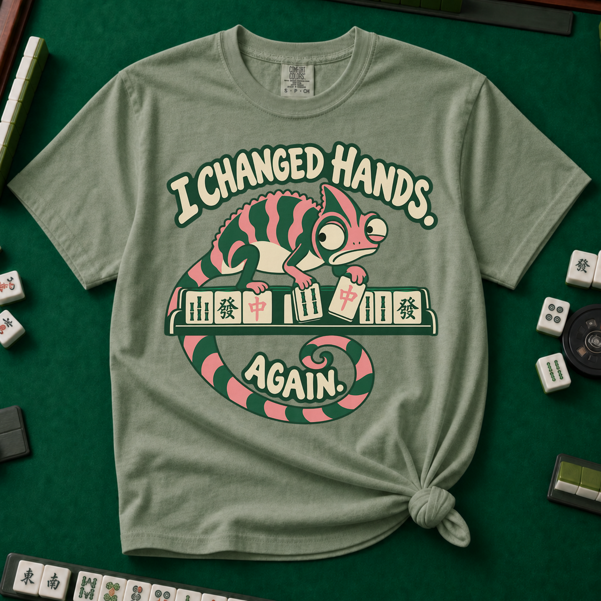 I Changed Hands Again T-Shirt in Moss - knotted tee on a green mahjong mat with scattered tiles, printed with a chameleon standing on a rack of mahjong tiles holding a red dragon tile, and the words I Changed Hands Again