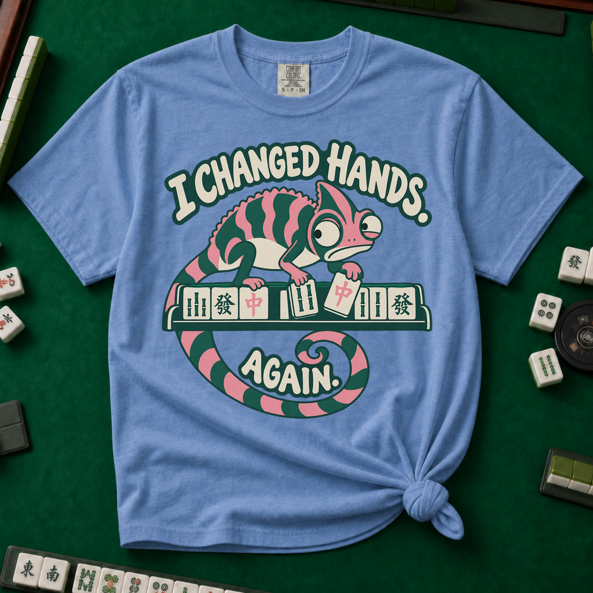 I Changed Hands Again T-Shirt in White - knotted tee on a green mahjong mat with scattered tiles, printed with a chameleon standing on a rack of mahjong tiles holding a red dragon tile, and the words I Changed Hands Again