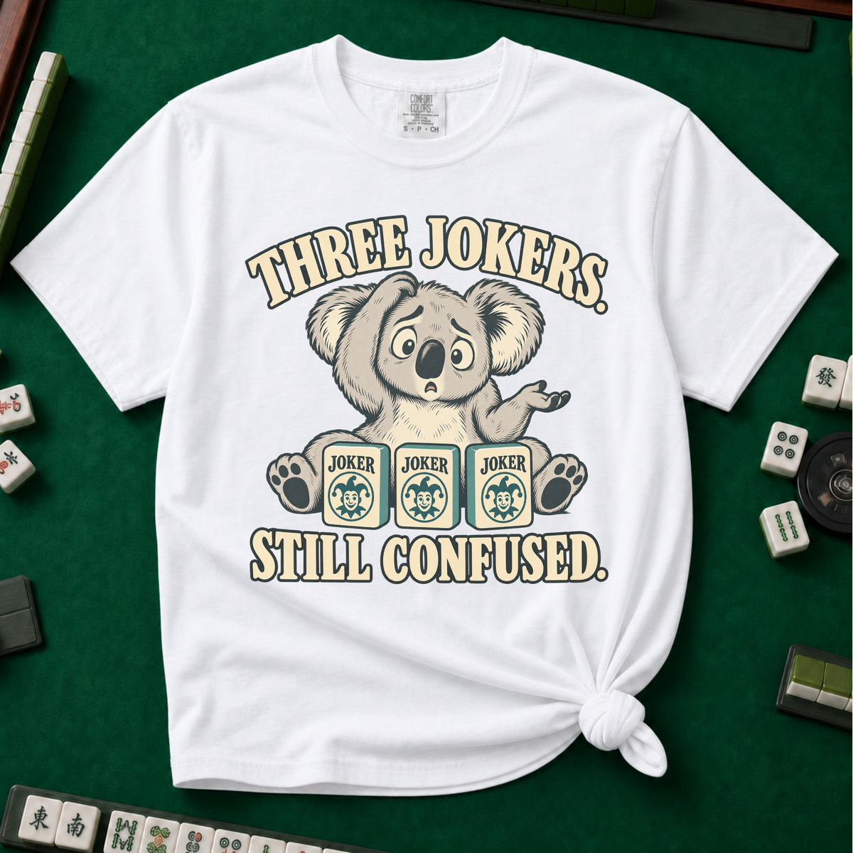 Three Jokers Still Confused T-Shirt in Blossom - knotted tee on a pale pink surface with mahjong tiles scattered around, printed with a confused koala shrugging behind three joker tiles, and the words Three Jokers Still Confused