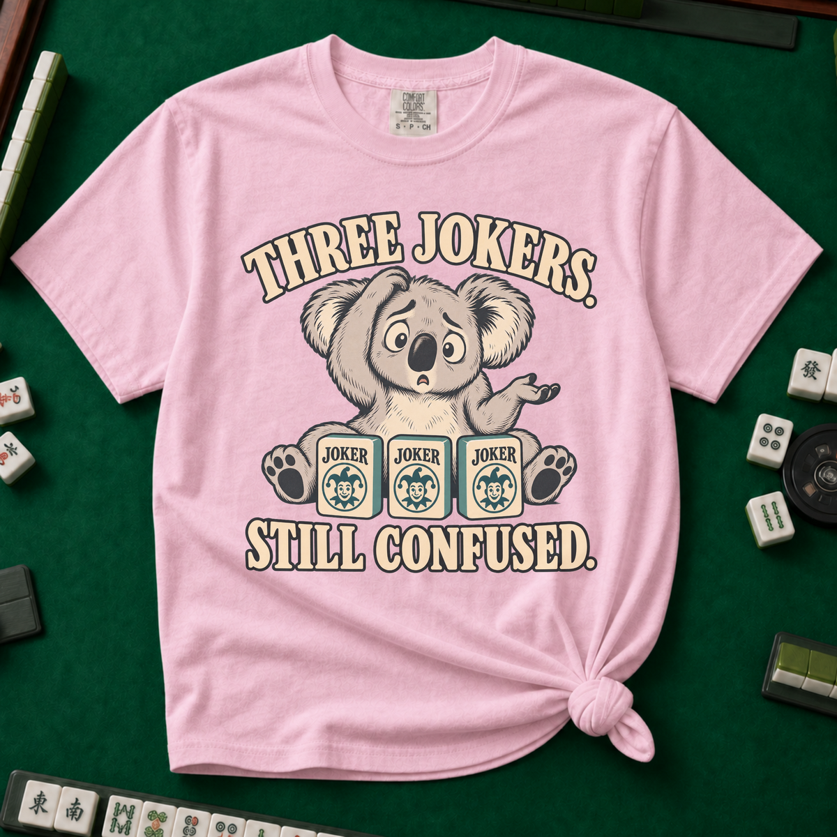 Three Jokers Still Confused T-Shirt in White - knotted tee on a green mahjong mat with scattered tiles, printed with a confused koala shrugging behind three joker tiles, and the words Three Jokers Still Confused