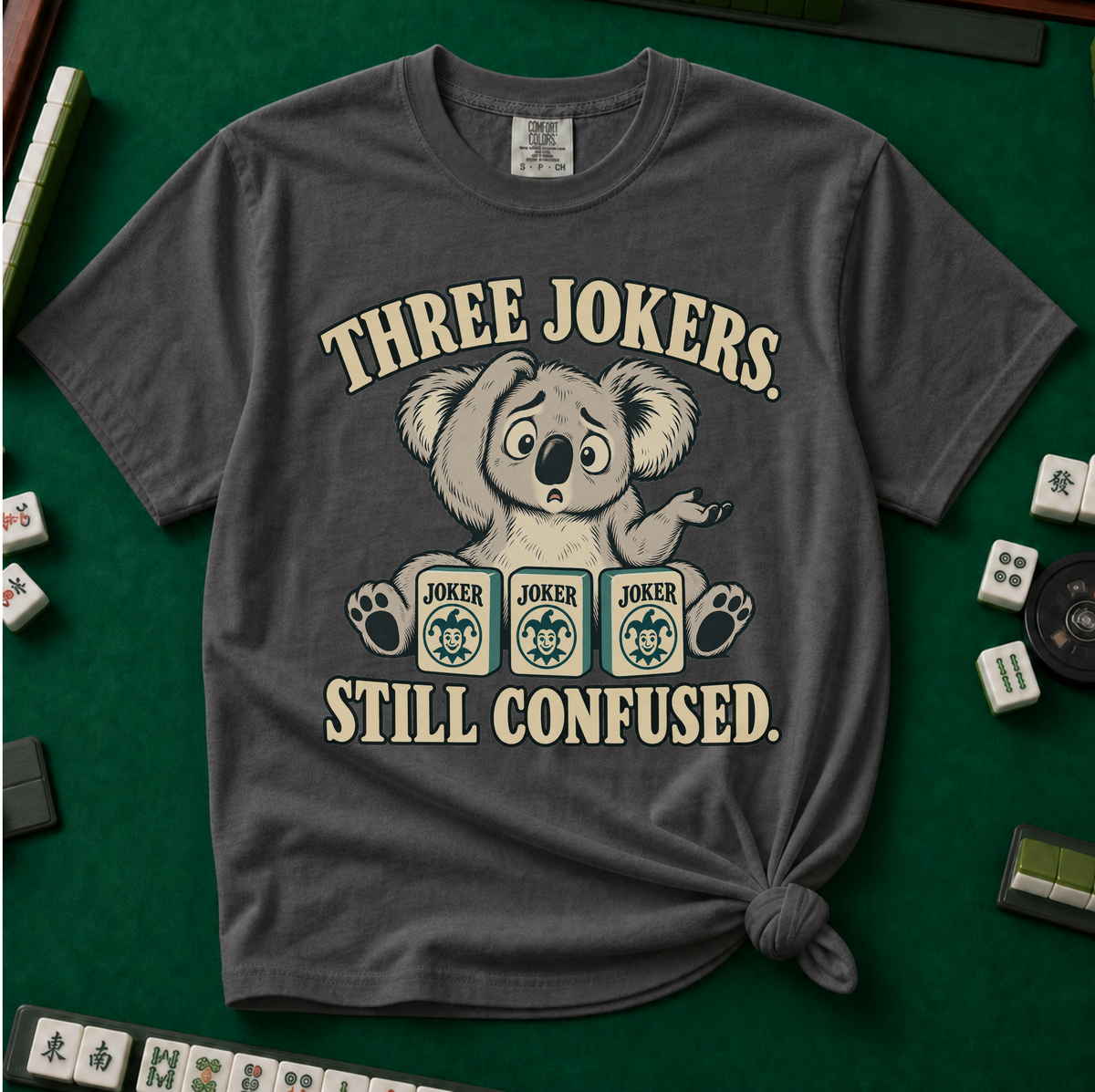 Three Jokers Still Confused T-Shirt in Moss - knotted tee on a green mahjong mat with scattered tiles, printed with a confused koala shrugging behind three joker tiles, and the words Three Jokers Still Confused