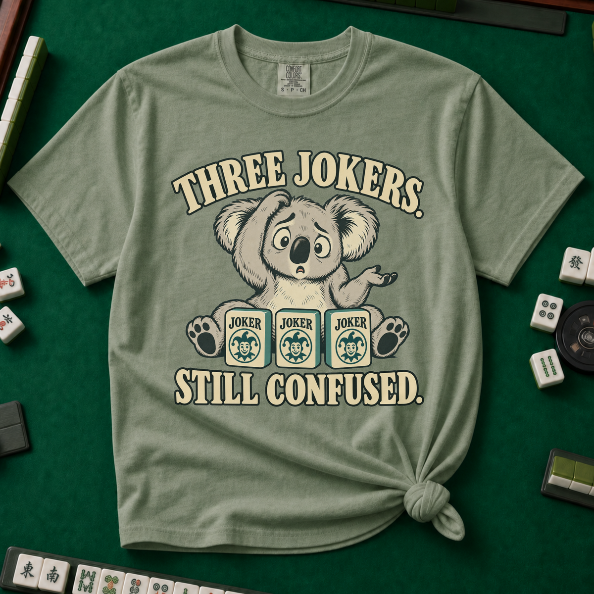 Three Jokers Still Confused T-Shirt in Espresso - knotted tee on a green mahjong mat with scattered tiles, printed with a confused koala shrugging behind three joker tiles, and the words Three Jokers Still Confused