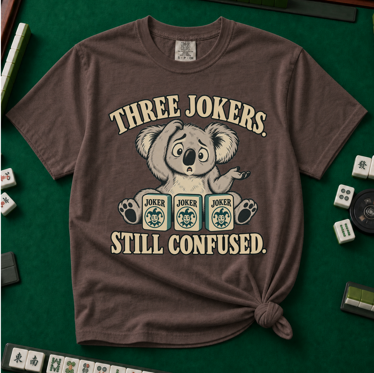 Three Jokers Still Confused T-Shirt in Pepper - knotted tee on a green mahjong mat with scattered tiles, printed with a confused koala shrugging behind three joker tiles, and the words Three Jokers Still Confused