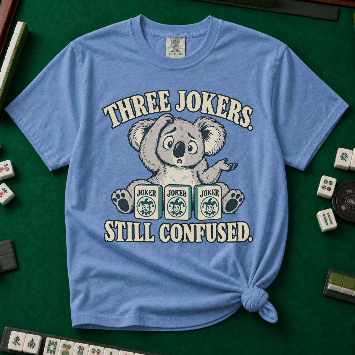 Three Jokers Still Confused T-Shirt in Black - knotted tee on a light surface with mahjong tiles scattered around, printed with a confused koala shrugging behind three joker tiles, and the words Three Jokers Still Confused