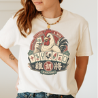 Cluck And Win Mahjong Rooster T-Shirt in Ivory - tee laid flat on a green mahjong mat with scattered tiles, printed with a rooster holding a green dragon tile above a fan of mahjong tiles in a red Cluck and Win badge