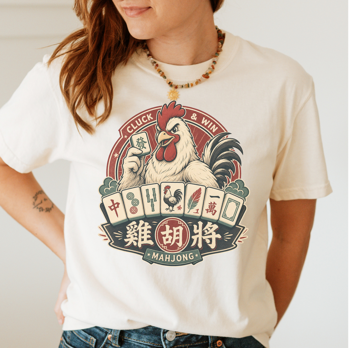 Cluck And Win Mahjong Rooster T-Shirt in Ivory - tee laid flat on a green mahjong mat with scattered tiles, printed with a rooster holding a green dragon tile above a fan of mahjong tiles in a red Cluck and Win badge