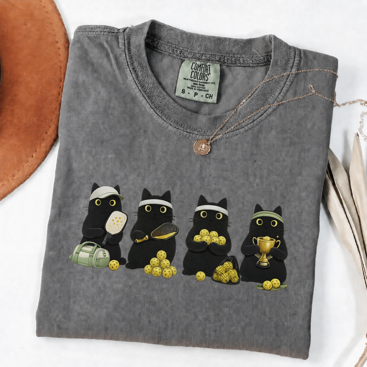 Four black cats with pickleball paddles, balls and a trophy printed on a Pepper Comfort Colors T-shirt.