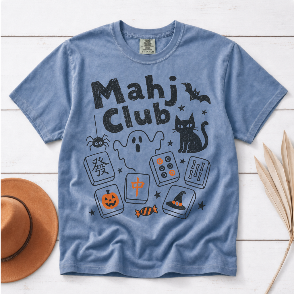 A light blue short-sleeve t-shirt with a 'Mahj Club' Halloween graphic, styled flat on a white wood background with a brown hat and dried palm leaves.