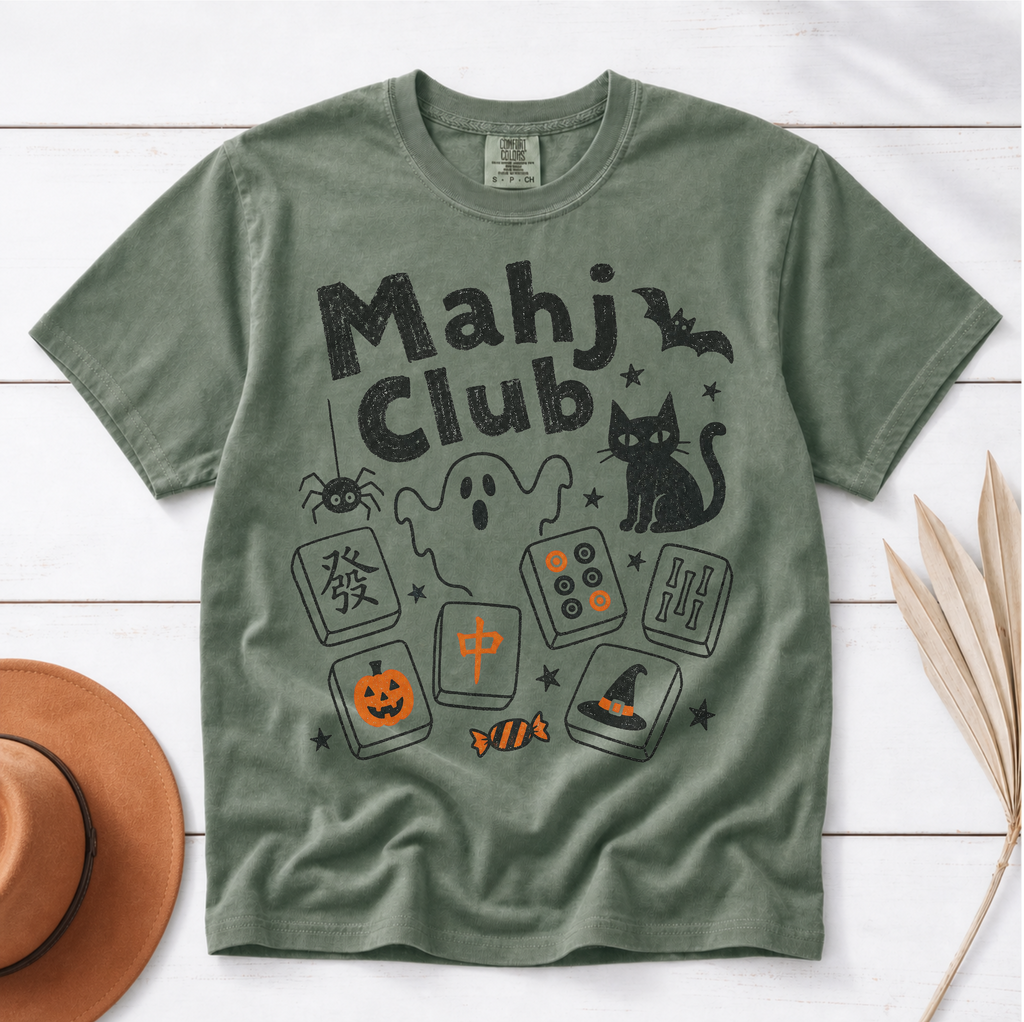 An olive green short-sleeve t-shirt with a 'Mahj Club' Halloween graphic, styled flat on a white wood background with a brown hat and dried palm leaves.