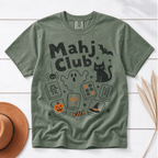 An olive green short-sleeve t-shirt with a 'Mahj Club' Halloween graphic, styled flat on a white wood background with a brown hat and dried palm leaves.