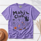 A purple short-sleeve t-shirt with a 'Mahj Club' Halloween graphic, laid flat on a white wood background with a brown hat and dried palm leaves as props.