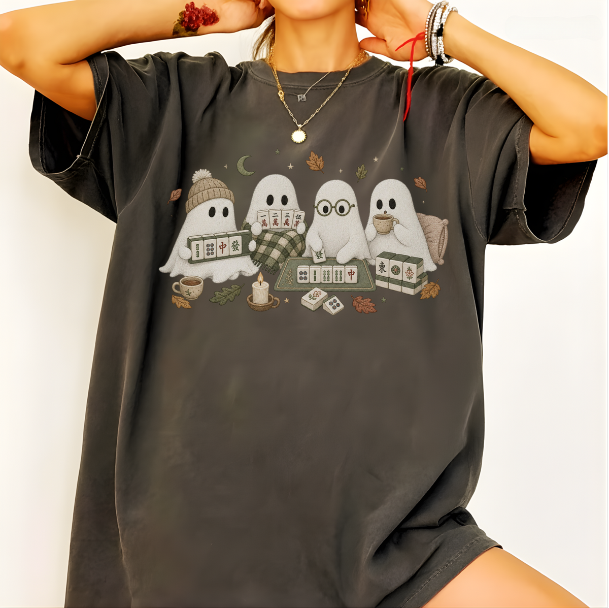A person wearing an oversized dark charcoal gray graphic T-shirt. The shirt features a cute illustration of four white cartoon ghosts sitting together on a blanket, surrounded by cozy autumn elements like a mug, coffee cups, cards, and leaves.
