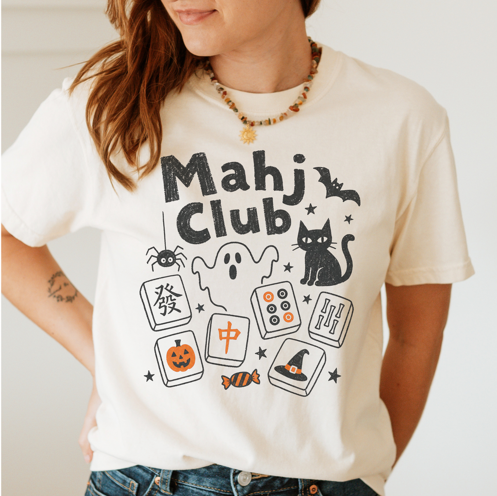 A woman wearing a cream-colored t-shirt with a black and orange 'Mahj Club' graphic featuring Halloween-themed illustrations like a ghost, cat, spider, and jack-o'-lantern.