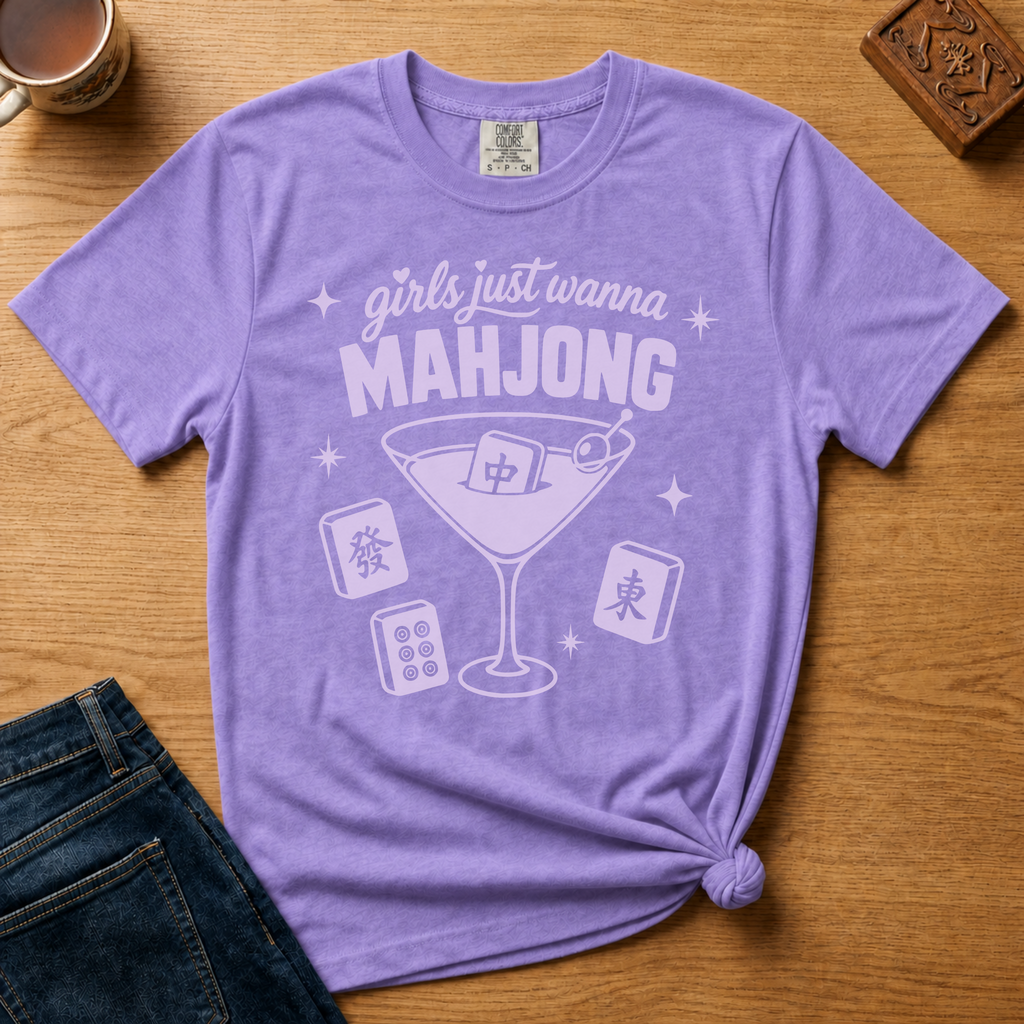 Girls Just Wanna Mahjong Shirt in Violet - pink lettering and a martini glass with a mahjong tile on a folded purple Comfort Colors 1717 tee, styled flat lay