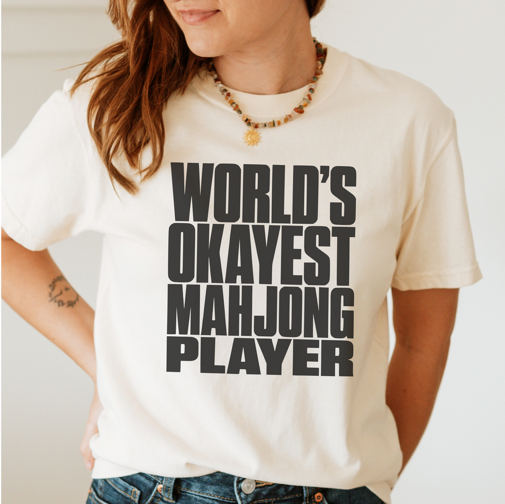 A woman wearing a cream-colored graphic t-shirt with bold black text that reads 'WORLD'S OKAYEST MAHJONG PLAYER'. The photo is cropped from the chin down to the waist, showing the fit of the shirt and the graphic print.