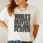 A woman wearing a cream-colored graphic t-shirt with bold black text that reads 'WORLD'S OKAYEST MAHJONG PLAYER'. The photo is cropped from the chin down to the waist, showing the fit of the shirt and the graphic print.
