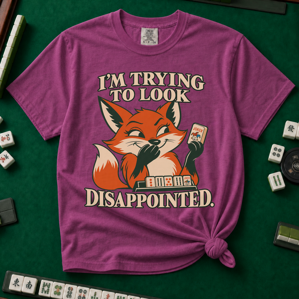I'm Trying To Look Disappointed T-Shirt in Berry, knotted flat lay on a green mahjong mat - smug retro fox holding up a joker tile, hiding a grin behind one paw, tile rack in front - "I'M TRYING TO LOOK DISAPPOINTED." in cream collegiate letters