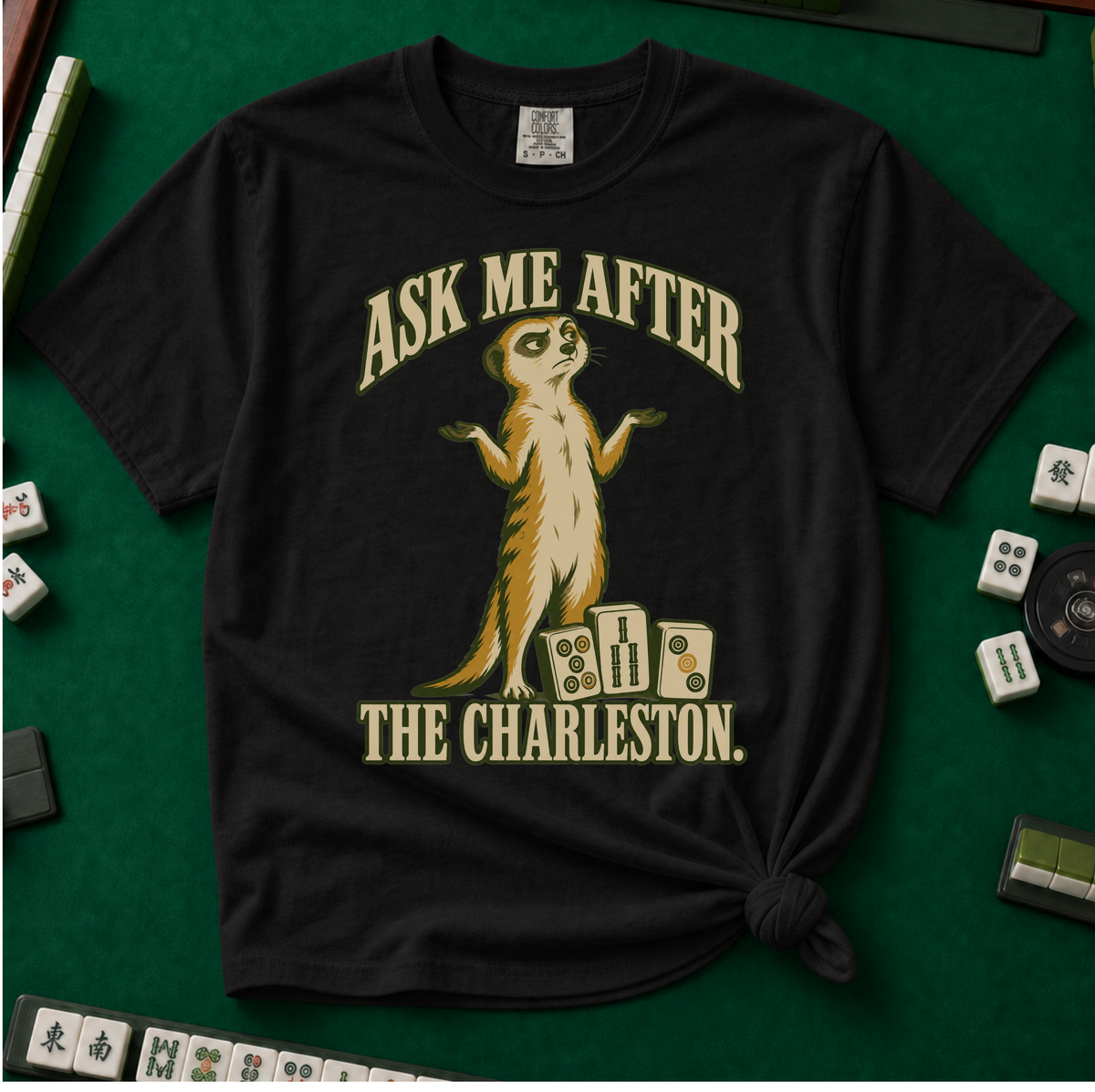 Ask Me After The Charleston T-Shirt in Black, knotted flat lay on a green mahjong mat - shrugging meerkat standing beside three mahjong tiles - "ASK ME AFTER THE CHARLESTON." in cream collegiate letters
