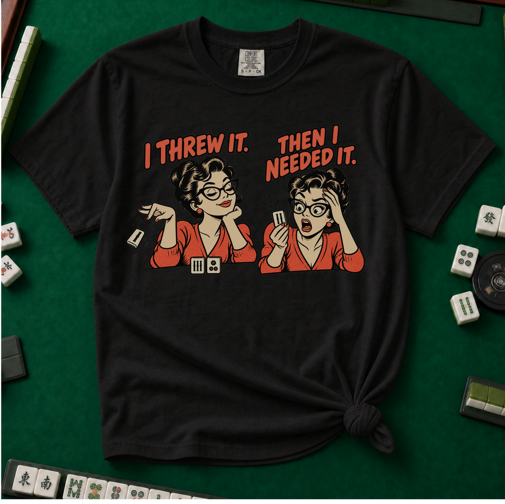 I Threw It Then I Needed It T-Shirt in Black, knotted flat lay on a green mahjong mat - a retro comic two-panel scene of a woman smugly flicking away a mahjong tile, then panicking when she needs it, with the words "I THREW IT. THEN I NEEDED IT."
