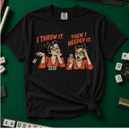 I Threw It Then I Needed It T-Shirt in Black, knotted flat lay on a green mahjong mat - a retro comic two-panel scene of a woman smugly flicking away a mahjong tile, then panicking when she needs it, with the words "I THREW IT. THEN I NEEDED IT."