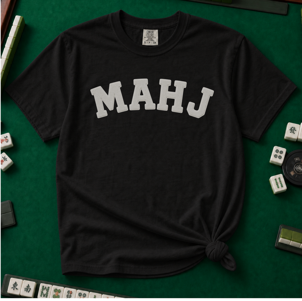 MAHJ Varsity T-Shirt in Black, knotted flat lay on a green mahjong mat - "MAHJ" in big white varsity arch letters with a stitched letterman look