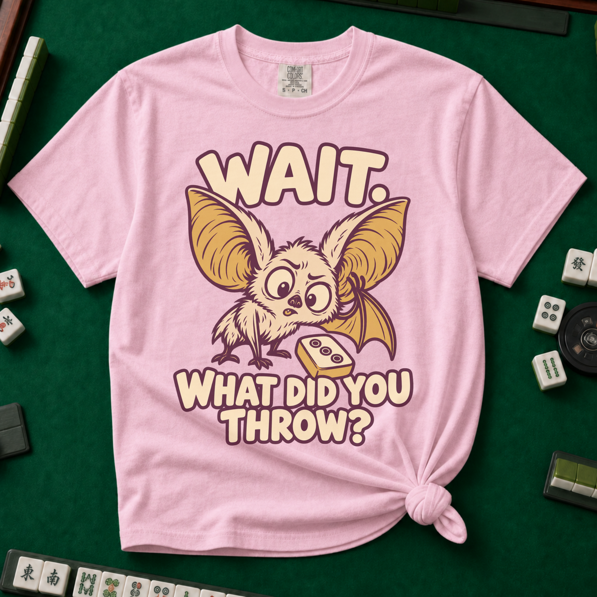 Wait What Did You Throw T-Shirt in Blossom, knotted flat lay on a green mahjong mat - a wide-eyed fluffy bat with huge golden ears leaning in over a fallen dot tile - "WAIT. WHAT DID YOU THROW?" in cream bubble letters