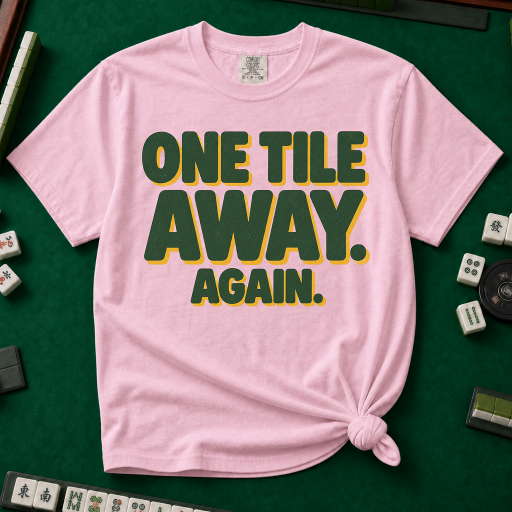 One Tile Away Again T-Shirt in Blossom, knotted flat lay on a green mahjong mat - "ONE TILE AWAY. AGAIN." in bold deep-green block letters with a gold drop shadow